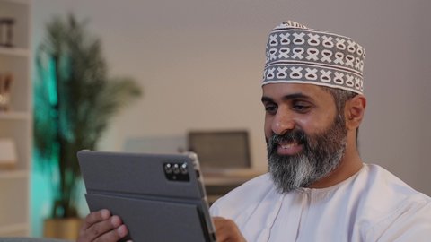 النقر على شاشة التابلت, ايماءات السعادة و السرور، رجل شايب عربي خليجي عماني مبتسم يرتدي الدشداشة و الكمة يمسك بيده الجهاز اللوحي, تصفح مواقع التواصل الاجتماعي, استخدام الاجهزة التقنية الحديثة
