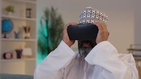 محاكاة العالم الخيالي، المتعة و التسلية بالمشاهدة عبر نظارات الواقع الافتراضي، استخدام جهاز تقني حديث و متطور،  رجل شايب عربي خليجي عماني مبتسم يرتدي الدشداشة و الكمة و يضع نظارة الفي ار ثلاثية الأبعاد
