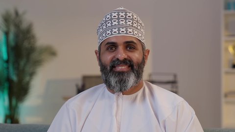 النظر الى الكاميرا بايماءات البهجة لامتلاك سكن حديث, تسويق المنازل و العقارات،  رجل شايب عربي خليجي عماني مبتسم يرتدي الدشداشة و الكمة يحمل بيده مجسم صغير خشبي لمنزل, مفهوم الاسكان و التمليك