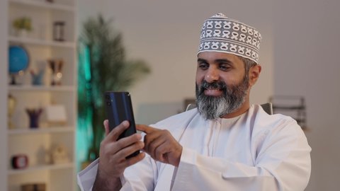 الدردشة عبر الانترنت, استخدام التقنية الحديثة المتطورة في الحياة اليومية, ايماءات السعادة و السرور، رجل شايب عربي خليجي عماني مبتسم يرتدي الدشداشة و الكمة يمسك بيده موبايل, تصفح مواقع التواصل الاجتماعي