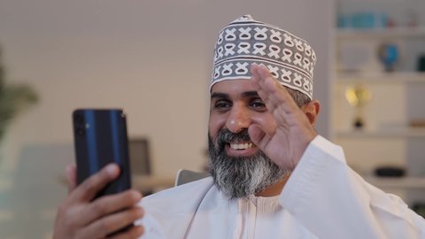 رفع كف الايد بايماءات السلام و التحية، استخدام التقنية الحديثه المتطورة في الحياة العملية, ايماءات السعادة و السرور، رجل شايب عربي خليجي عماني مبتسم يرتدي الدشداشة و الكمة يمسك بيده موبايل, اجراء مكالمة فيديو عبر الانترنت