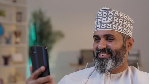 اجراء مكالمة فيديو عبر الانترنت، استخدام الاجهزة التقنية الحديثة، رفع كف الايد بايماءات السلام و التحية, ايماءات السعادة و السرور، رجل شايب عربي خليجي عماني مبتسم يرتدي الدشداشة و الكمة يمسك بيده الموبايل و يرفع اصبع الابهام باشارة اللايك