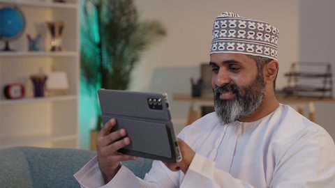 النقر على شاشة الجهاز اللوحي، استخدام اجهزة تقنية حديثة، مفهوم إدارة المشاريع عن بعد، ايماءات السعادة و السرور، رجل شايب عربي خليجي عماني مبتسم يرتدي الدشداشة و الكمة يمسك بيده التابلت, تصفح مواقع التواصل الاجتماعي