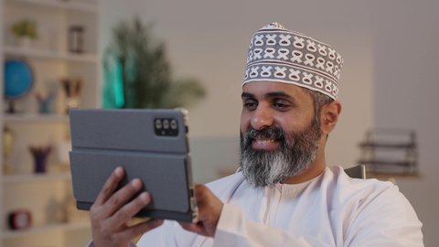 النقر بالاصبع على الشاشة، استخدام اجهزة تقنية حديثة،  مفهوم إدارة المشاريع عن بعد، ايماءات السعادة و السرور، رجل شايب عربي خليجي عماني مبتسم يرتدي الدشداشة و الكمة يمسك بيده التابلت, تصفح مواقع التواصل الاجتماعي
