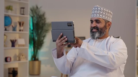 دمج التقنية و التكنولوجيا في الروتين اليومي، مفهوم العمل عن بعد، ايماءات التركيز والسرور، قضاء وقت ممتع،  رجل شايب عربي خليجي عماني مبتسم يرتدي الدشداشة و الكمة يمسك بيده التابلت, تصفح مواقع التواصل الاجتماعي