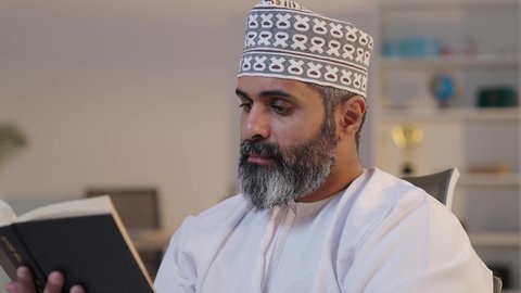 The culture of self-learning, harmony, tranquility, and serenity, enjoying reading a book, an elderly Arab Gulf Omani man smiling, wearing a dishdasha and a kumma, holding a book in his hand, gestures of focus and immersion, having a good time.