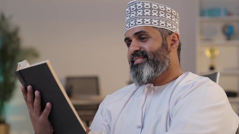 قراءة الروايات و القصص, ثقافة التعلم الذاتي, الانسجام و الهدوء و السكينة,  رجل شايب عربي خليجي عماني مبتسم يرتدي الدشداشة و الكمة يمسك بيده كتاب,  إيماءات التركيز و الاندماج, قضاء وقت ممتع
