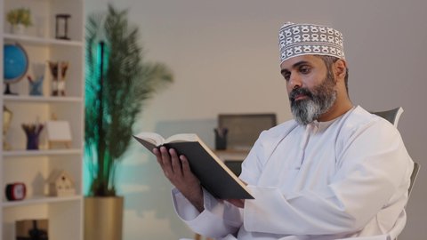 الانسجام و الهدوء و السكينة, الاستمتاع بقراءة كتاب،  رجل شايب عربي خليجي عماني مبتسم يرتدي الدشداشة و الكمة يمسك بيده كتاب,  إيماءات التركيز و الاندماج, قضاء وقت ممتع