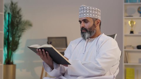قراءة الكتب والقصص و الروايات, ثقافة التعلم الذاتي,  رجل شايب عربي خليجي عماني مبتسم يرتدي الدشداشة و الكمة يمسك بيده كتاب,  إيماءات التركيز و الاندماج, قضاء وقت ممتع