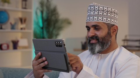 تصفح مواقع التواصل الاجتماعي، ايماءات السعادة و السرور، استخدام الاجهزة التقنية الحديثة، مفهوم إدارة المشاريع عن بعد، رجل شايب عربي خليجي عماني مبتسم يرتدي الدشداشة و الكمة يمسك بيده التابلت