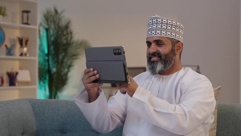 مفهوم إدارة المشاريع عن بعد، ايماءات السعادة و السرور، رجل شايب عربي خليجي عماني مبتسم يرتدي الدشداشة و الكمة يمسك بيده التابلت, تصفح مواقع التواصل الاجتماعي, استخدام الاجهزة التقنية الحديثة