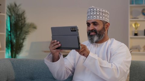 الاحترافية و التفاني في الإنجاز، ايماءات السعادة و السرور، استخدام الاجهزة التقنية الحديثة، مفهوم إدارة المشاريع عن بعد، رجل شايب عربي خليجي عماني مبتسم يرتدي الدشداشة و الكمة  يمسك بيده التابلت