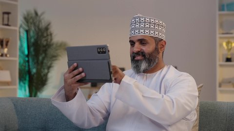 استخدام الاجهزة التقنية الحديثة، مفهوم إدارة المشاريع عن بعد، ايماءات السعادة و السرور، رجل شايب عربي خليجي عماني مبتسم يرتدي الدشداشة و الكمة  يمسك بيده التابلت, تصفح مواقع التواصل الاجتماعي