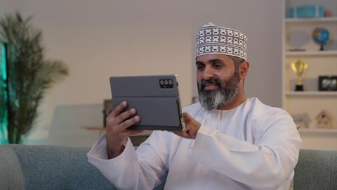 ايماءات السعادة و السرور، استخدام الاجهزة التقنية الحديثة، مفهوم إدارة المشاريع عن بعد، تصفح مواقع التواصل الاجتماعي، رجل شايب عربي خليجي عماني مبتسم يرتدي الدشداشة و الكمة يمسك بيده التابلت
