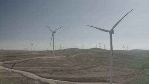 توليد الكهرباء بواسطة مزارع الرياح، مناخ قاسٍ وجاف، مفهوم استخدام الطاقات المتجددة صديقة البيئة، التنمية المستدامة بالطاقة البديلة في دومه الجندل بالمملكة العربية السعودية، تصوير درون جوي لحقول توربينات الرياح وطواحين الهواء وسط الرمال الذهبية في المناطق الصحرواية