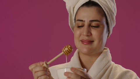 اتباع نظام غذائي صحي، استخدام عسل النحل والمنتجات الطبيعية للعناية بالبشرة، فيتامينات مغذية للجسم، منتج وطني محلي سعودي، بورتريه لامراة عربية خليجية سعودية ترتدي روب استحمام ومنشفة بيضاء تحمل ملعقة خشبية و تنظر الى الكاميرا بسعادة و سرور, خلفية بنفسجية