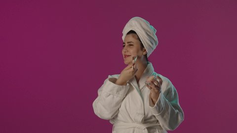 اخفاء عيوب البشرة, الاهتمام و العناية بالجمال, الشغف بوضع المكياج, بورترية لمرأة عربية خليجية سعودية مبتسمة ترتدي روب حمام و منشفة بيضاء و تستخدم الفرشاة و كريم الأساس, خلفية بنفسجية