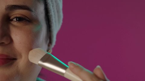 الاهتمام بالأناقة و المظهر الخارجي، مفهوم التزين و التجمل, الشغف بوضع المكياج, بورترية لمرأة عربية خليجية سعودية مبتسمة ترتدي منشفة بيضاء و تستخدم الفرشاة و كريم الأساس, خلفية بنفسجية