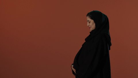 الاهتمام بصحة الجنين، مشاعر الامومة و حضن طفلها، مفهوم الحمل والانجاب، بورتريه لإمرأة عربية خليجية سعودية محجبة ترتدي عباءة سوداء تضع يدها على بطنها بايماءات الحماس, خلفية بني