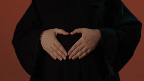 مشاعر الامومة و حضن طفلها، مفهوم الحمل والانجاب، الاهتمام بصحة الجنين،  بورتريه لإمرأة عربية خليجية سعودية محجبة ترتدي عباءة سوداء تضع يدها على بطنها بايماءات الحماس, خلفية بني