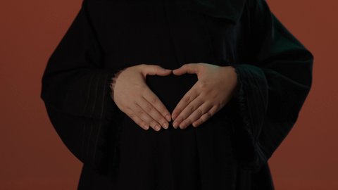 ايماءات السعادة و الحماس, الحفاظ على صحة الأم و الطفل، مشاعر الامومة و حضن طفلها، مفهوم الحمل والانجاب، الاهتمام بصحة الجنين، بورتريه لإمرأة عربية خليجية سعودية محجبة ترتدي عباءة سوداء تضع يدها على بطنها, خلفية بني