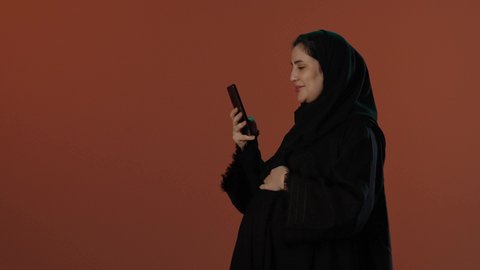 استخدام الهاتف المحمول و الأجهزة التقنية الحديثة،  الاهتمام بصحة الجنين، مفهوم الحمل والانجاب، بورتريه لإمرأة عربية خليجية سعودية محجبة ترتدي عباءة سوداء تمسك الموبايل و تضع يدها على بطنها تنتظر قدوم مولودها, خلفية بني