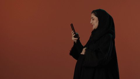Following social media news using a mobile phone, caring for the health of the fetus, the concept of pregnancy and childbirth, a portrait of a Saudi Arabian Gulf woman wearing a black abaya, holding a mobile phone and looking at the camera with gestures of happiness, brown background.