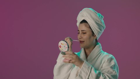 الاستعانة بالمرآة في وضع المكياج, استعمال المستحضرات التجميلية, مفهوم التزين و التجمل, العناية بالجمال و المظهر الخارجي, بورترية لمرأة عربية خليجية سعودية  ترتدي روب حمام و منشفة بيضاء و تستخدم أحمر الشفاه, خلفية بنفسجية