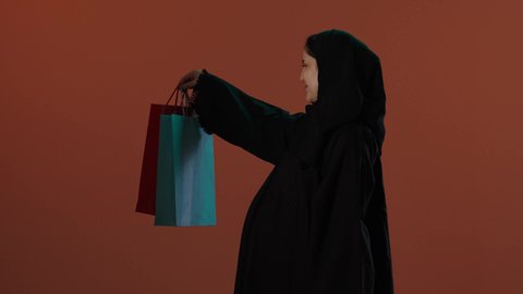 الاستفادة من العروض الموسمية و التخفيضات، الأثر الايجابي للتسوق على نفسية المرأة، مفهوم الحمل والانجاب، بورتريه لإمرأة عربية خليجية سعودية محجبة ترتدي عباءة سوداء تحمل بيدها أكياس التسوق الملونة، خلفية بني