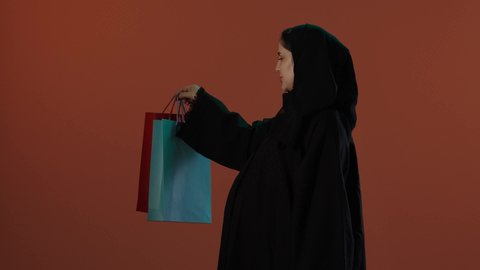 الاستفادة من العروض والتخفيضات، الأثر الايجابي للتسوق على نفسية المرأة، مفهوم الحمل والانجاب، بورتريه لإمرأة عربية خليجية سعودية محجبة ترتدي عباءة سوداء تحمل بيدها أكياس التسوق الملونة، خلفية بني