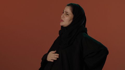 وجع في الظهر و العمود الفقري خلال الحمل، الاهتمام بصحة الجنين، الحفاظ على صحة الأم و الطفل، الأمومة و بداية حياة جديدة، بورتريه لإمرأة عربية خليجية سعودية محجبة ترتدي عباءة سوداء تضع يدها على بطنها بايماءات التألم, خلفية بني