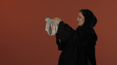 الاستعداد لاستقبال المولود الجديد, اختيار ملابس مواليد رضع، مفهوم الحمل والانجاب،  بورتريه لإمرأة عربية خليجية سعودية محجبة ترتدي عباءة سوداء تمسك بيدها افرول، خلفية بني