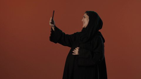مشاركة الأخبار السعيدة مع الأهل و الأصدقاء, أهمية التكنولوجية في التواصل, مفهوم الحمل والانجاب،  بورتريه لإمرأة عربية خليجية سعودية محجبة ترتدي عباءة سوداء تقوم باجراء مكالمة فيديو عبر الهاتف المحمول، خلفية بني