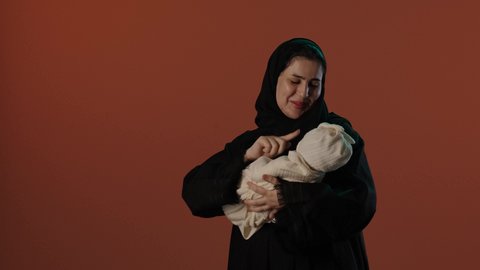 مفهوم الأمومة و الطفولة, تحمل المسؤولية والعناية بالأطفال، بورتريه لإمرأة عربية خليجية سعودية مبتسمة محجبة  ترتدي عباءة سوداء تداعب صغيرها بإيماءات الحب والحنان, خلفية بني