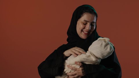 ايماءات الحب و الحنان, الأمومة و حماية الأطفال و رعايتهم,  بورتريه لإمرأة عربية خليجية سعودية محجبة ترتدي عباءة سوداء تداعب صغيرها, النظر الى الكاميرا بتعابير السعادة و السرور,  خلفية بني