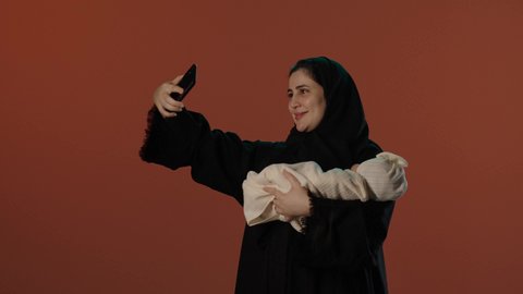 استخدام الأجهزة و التقنيات الحديثة، ايماءات الحب و الحنان, الأمومة و حماية الأطفال و رعايتهم, بورتريه لامرأة عربية خليجية سعودية محجبة ترتدي عباءة سوداء تقوم باجراء مكالمة فيديو عبر الهاتف المحمول، خلفية بني