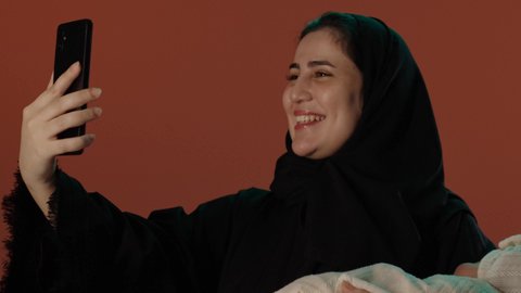 التواصل مع الأهل و الأصدقاء، استخدام الأجهزة و التقنيات الحديثة، ايماءات الحب و الحنان, الأمومة و حماية الأطفال و رعايتهم,  بورتريه لامرأة عربية خليجية سعودية محجبة ترتدي عباءة سوداء تقوم باجراء مكالمة فيديو عبر الهاتف المحمول، خلفية بني