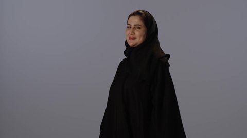تعابير الثقة و الاعتزاز بالنفس, مفهوم الانوثة و النعومة, النظر إلى الكاميرا بإيماءات السعادة والسرور, بورتريه لامرأة عربية خليجية سعودية محجبة ترتدي عباءة سوداء تقف باستقامة و اعتدال و أيدي متقاطعة, خلفية رمادية