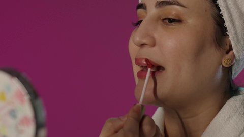 استعمال المستحضرات التجميلية, مفهوم التزين و التجمل, العناية بالجمال و المظهر الخارجي, بورترية لمرأة عربية خليجية سعودية تستخدم أحمر الشفاه و تنظر في المرآة، العناية بالنفس و الاطلالة المتألقة, ايماءات السرور و السعادة، خلفية بنفسجية