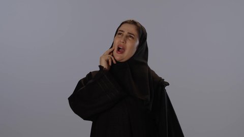 الحاجة للعلاج و الذهاب للعيادة السنية, الشعور بالتعب والمرض, الإحساس بوجع الأسنان و النخور،  بورتريه لامرأة عربيه خليجيه سعودية محجبة ترتدي عباءة سوداء تضع يدها على خدها بتعابير الألم, خلفية رمادية