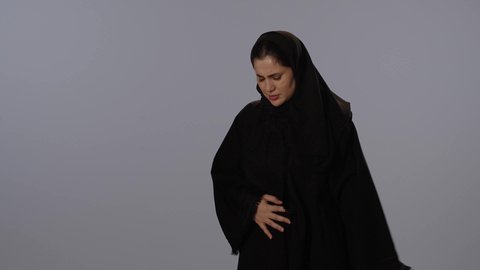 عسر الهضم و الشعور بحرقة في المعدة، إيماءات الألم و المعاناة،  بورتريه لإمرأة عربية خليجية سعودية محجبة ترتدي عباءة سوداء تضع يدها على بطنها بتعابير التألم، مفهوم التعبير بالإشارة, خلفية رمادية