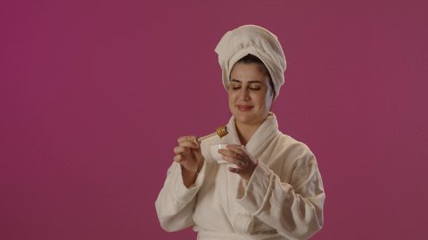 استخدام المنتجات الطبيعية للعناية بالبشرة، ايماءات السرور و السعادة، اتباع حمية غذائية، منتج وطني محلي سعودي، بورتريه لمرأة عربية خليجية سعودية  ترتدي روب حمام و منشفة بيضاء تحمل بيدها ملعقة خشبية مغمورة بالعسل، خلفية بنفسجية