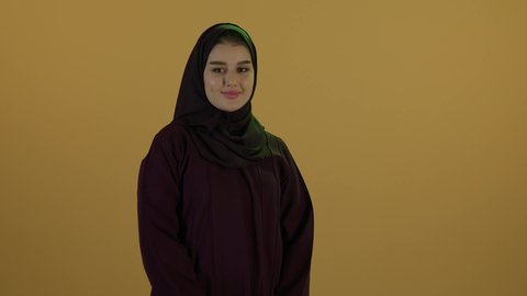 النظر الى الكاميرا بايماءات السرور، مفهوم الحمل والانجاب، الاهتمام بصحة الجنين، الحفاظ على صحتها و صحة طفلها، إمرأة عربية خليجية سعودية تمسك بيدها حذاء أطفال، تصوير مقرب لإمراة عربية خليجة سعودية ترتدي العباءة والحجاب تقف بايدي متقاطعة، خلفية بيج