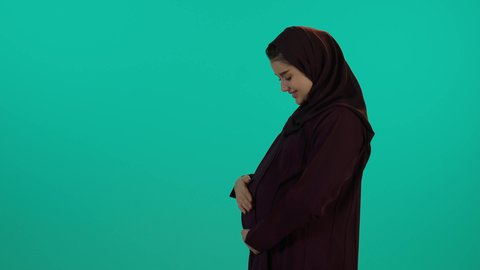الحفاظ على صحتها و صحة طفلها، وضع اليد على بطنها تنتظر قدوم مولودها، مفهوم الحمل والانجاب، الاهتمام بصحة الجنين، الحفاظ على صحتها و صحة طفلها، الاهتمام بصحة الجنين،  تصوير مقرب لإمراة عربية خليجة سعودية ترتدي العباءة والحجاب  بتعابير السرور، خلفية زرقاء