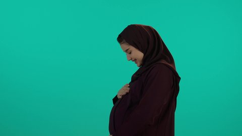 وضع اليد على بطنها تنتظر قدوم مولودها، الحفاظ على صحتها و صحة طفلها،  مفهوم الحمل والانجاب، الاهتمام بصحة الجنين، الحفاظ على صحتها و صحة طفلها، الاهتمام بصحة الجنين،  تصوير مقرب لإمراة عربية خليجة سعودية ترتدي العباءة والحجاب  بتعابير السرور، خلفية زرقاء