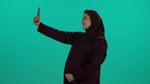 استخدام الهاتف المحمول في الحياة اليومية، توثيق لحظات الحمل السعيدة بصورة سيلفي، مفهوم الحمل والانجاب، الاهتمام بصحة الجنين، الحفاظ على صحتها و صحة طفلها، الاهتمام بصحة الجنين،  تصوير مقرب لإمراة عربية خليجة سعودية ترتدي العباءة والحجاب  بتعابير السرور، خلفية زرقاء