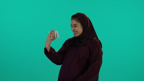 تأمل صورة سونار لجنين باللون الابيض والاسود ومشاعر الامومة، مفهوم الحمل والانجاب، الاهتمام بصحة الجنين، الحفاظ على صحتها و صحة طفلها، الاستفادة من العروض الموسمية والتخفيضات، تصوير مقرب لإمراة عربية خليجة سعودية ترتدي العباءة والحجاب  بتعابير السرور، خلفية زرقاء