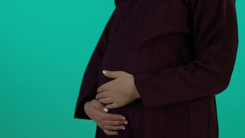 Affection and touching a pregnant woman's belly, the concept of pregnancy and childbirth, caring for the fetus's health, maintaining her health and the health of her child, taking advantage of seasonal offers and discounts, a close-up photograph of a Saudi Arabian woman wearing an abaya and hijab with expressions of joy, blue background