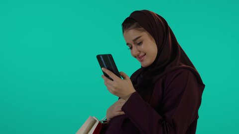 استخدام الهاتف لشراء للاطفال بتعابير السرور، مفهوم الحمل والانجاب، الاهتمام بصحة الجنين، الحفاظ على صحتها و صحة طفلها، الاستفادة من العروض الموسمية والتخفيضات، تصوير مقرب لإمراة عربية خليجة سعودية ترتدي العباءة والحجاب تحمل أكياس تسوق ملونة، خلفية زرقاء
