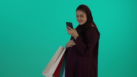 الاطلاع على مواقع الشراء للاطفال بتعابير السرور، مفهوم الحمل والانجاب، الاهتمام بصحة الجنين، الحفاظ على صحتها و صحة طفلها، الاستفادة من العروض الموسمية والتخفيضات، تصوير مقرب لإمراة عربية خليجة سعودية ترتدي العباءة والحجاب تحمل أكياس تسوق ملونة، خلفية زرقاء